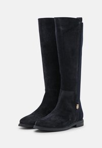 Bottes en daim noires jusqu'aux genoux avec une texture lisse, bout arrondi et accents en métal doré sur le côté. Panneaux élastiques latéraux pour un ajustement parfait.