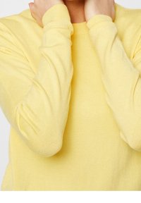 Gul stickad tröja med långa ärmar, med ribbad textur och manchetter. Materialet verkar mjukt och lätt.