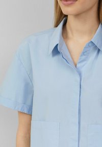 Camicetta azzurra chiara a maniche corte realizzata in tessuto morbido. Presenta un colletto, bottoni frontali e due tasche applicate. Design semplice.