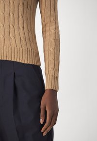 Gebreide kabeltrui in beige met geribbelde manchetten, gecombineerd met donkerblauwe broek. Close-up van de mouw en handdetail.