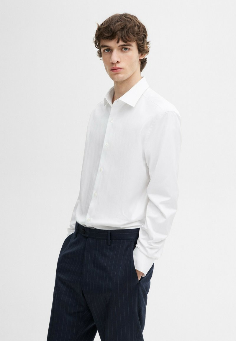 Jonge man met krullend haar, gekleed in een wit overhemd met knopen en marineblauwe pantalon met krijtstreep, staand met handen in de zakken.