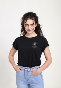Mladá žena s kudrnatými vlasy, oblečená v černém tričku s malým emblémem a v světlých modrých džínách, s rukama v kapsách, mírně se usmívající.