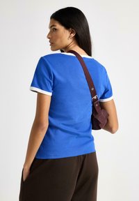 Blå bomulds t-shirt med hvid kant, tætsiddende design, korte ærmer, båret med mørkebrune bukser og en lille burgunderrød taske på en rem.