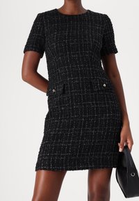 Svart tweedklänning med korta ärmar, en figurnära siluett, texturerat rutmönster, två framfickor och dekorativa hårdvarudetaljer.