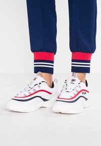 Zapatillas deportivas blancas con acentos en rojo y azul marino, que presentan una suela texturizada y un diseño de cordones. Mostradas con joggers azul marino y puños a rayas rojas.