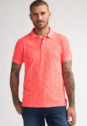Polo color coral con pequeños estampados en azul marino, mangas cortas y cuello con solapa. Combinado con vaqueros de mezclilla.