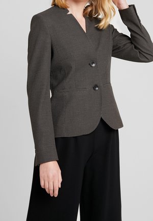 Vrouw draagt een nauwsluitend zwart-wit gestippeld colbert met twee knopen, over losse zwarte broek, hand naar het hoofd geheven.