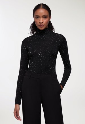 Maglia nera a maniche lunghe con colletto a dolcevita, caratterizzata da un motivo a pois argentati, realizzata in un materiale morbido, indossata con pantaloni neri a vita alta.