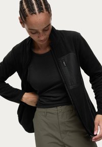 Chaqueta de forro polar negra con un bolsillo de pecho con cremallera, underneath una camiseta de tirantes negra acanalada, combinada con pantalones verde oliva claro. Textura suave.