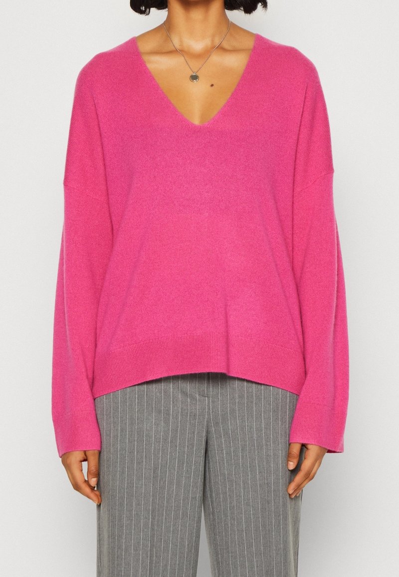 Suéter de gran tamaño color rosa brillante con escote en V, puños acanalados y dobladillo, combinado con pantalones de rayas grises, que presentan un corte relajado y una textura suave.