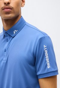 Άνδρας με μπλε κοντομάνικο πόλο πουκάμισο με το λογότυπο J.LINDEBERG στο γιακά και το μανίκι, φωτογραφημένος από το στήθος μέχρι το πηγούνι.