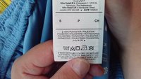 Vitt tygvårdsetikett med svart text som visar tvättanvisningar, material (100 % polyester) och plaggets storlek. Bakgrundstyget är ljusblått.