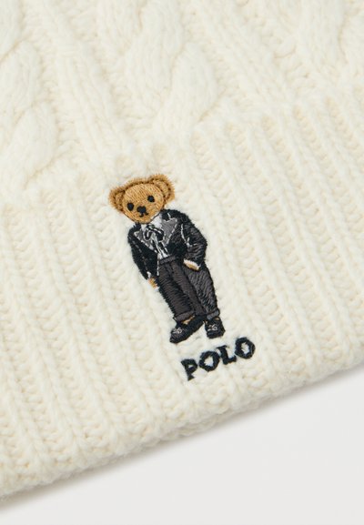 Sűrűn szőtt, krémszínű szövet, amelyen egy barna medve hímzés látható, fekete öltönyben és nyakkendőben, a "POLO" szó felett, fekete színben.