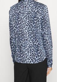 Blouse à imprimé léopard en bleu clair et bleu foncé avec manches longues, tissu lisse, ourlet arrondi et poignets froncés. Associée à un pantalon noir.