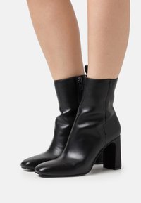 Schwarze Leder-Stiefeletten mit glatter Textur, spitzem Schuhkopf und Blockabsatz. Mit einem seitlichen Reißverschluss für einfaches An- und Ausziehen.