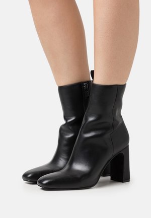 Schwarze Leder-Stiefeletten mit glatter Textur, spitzem Schuhkopf und Blockabsatz. Mit einem seitlichen Reißverschluss für einfaches An- und Ausziehen.