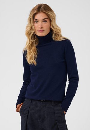 Femme blonde aux ondulations lâches portant un pull à col roulé bleu marine et un pantalon sombre à fines rayures, debout les mains dans les poches.