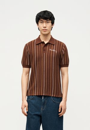 Giovane uomo che indossa una polo a maniche corte a righe marroni e jeans blu, in piedi davanti a uno sfondo chiaro e uniforme.