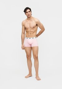 Νεαρός άνδρας που στέκεται ξυπόλητος, χωρίς πουκάμισο, φορώντας ανοιχτό ροζ μπόξερ με λευκή μέση που γράφει "GCDS" σε απλό φωτεινό φόντο.