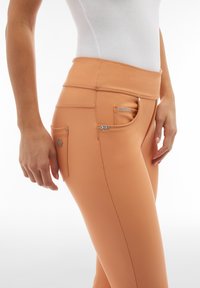 Leggings color pesca realizzati in tessuto elasticizzato, dotati di un'alta vita, cuciture decorative, tasca frontale e sottili accenti metallici.