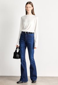 Sötétkék, szárában bővülő farmernadrág magas derékkal, fehér hosszú ujjú felsővel és kis fekete kézitáskával kombinálva. Fekete öv fém részletekkel.