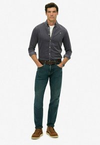 Man staat met zijn handen in zijn zakken, gekleed in een donkergrijze button-up shirt, blauwe jeans, een bruine riem, een horloge en bruine leren laarzen.