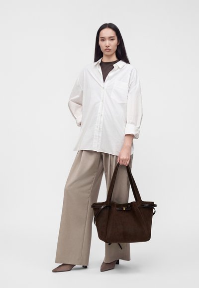 Femme portant une chemise blanche ample et un pantalon beige à jambes larges, tenant un grand sac cabas en suédine marron foncé avec de longues anses.
