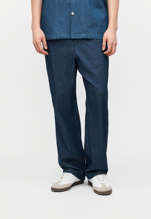 Henkilö, joka pukeutuu tummansinisiin leveälahkeisiin farkkuihin ja valkoisiin lenkkareihin, joissa on beigen sävyisiä yksityiskohtia, seisoo yksivärisen vaalean taustan edessä.