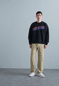 Schwarzer Hoodie mit blauem Aufdruck, beige lässige Hose mit Nahtdetails, weiße Sneakers, einfarbiger grauer Hintergrund.