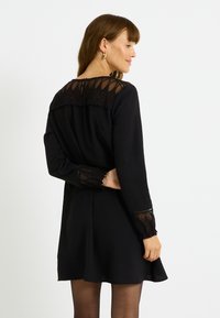 Robe noire à manches longues, présentant un empiècement en dentelle transparente et des détails aux poignets. Design de jupe évasée, fabriquée à partir d'un tissu doux.