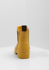 Botín de goma amarillo con una superficie texturizada, acentos marrones y una lengüeta. Presenta una punta redondeada y una suela resistente.