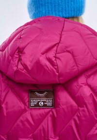 Fuchsiová péřová bunda s diamantovým prošíváním; obsahuje hnědou nášivku s textem. Doplňkem je modrý pletený čepice.