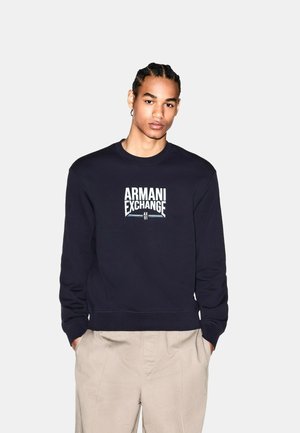 Giovane con capelli intrecciati che indossa una felpa nera Armani Exchange e pantaloni beige, in piedi con le mani nelle tasche davanti a uno sfondo semplice.