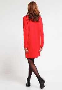 Rode jurk met lange mouwen, een rechte silhouet en een zachte textuur, met een achterste plooien en gedragen met zwarte enkelbooties.