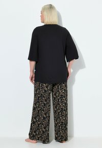 Zwart oversized top met korte mouwen, gecombineerd met wijde broekspijpen met een patroon van draaiende gouden en groene ontwerpen op een zwarte achtergrond.