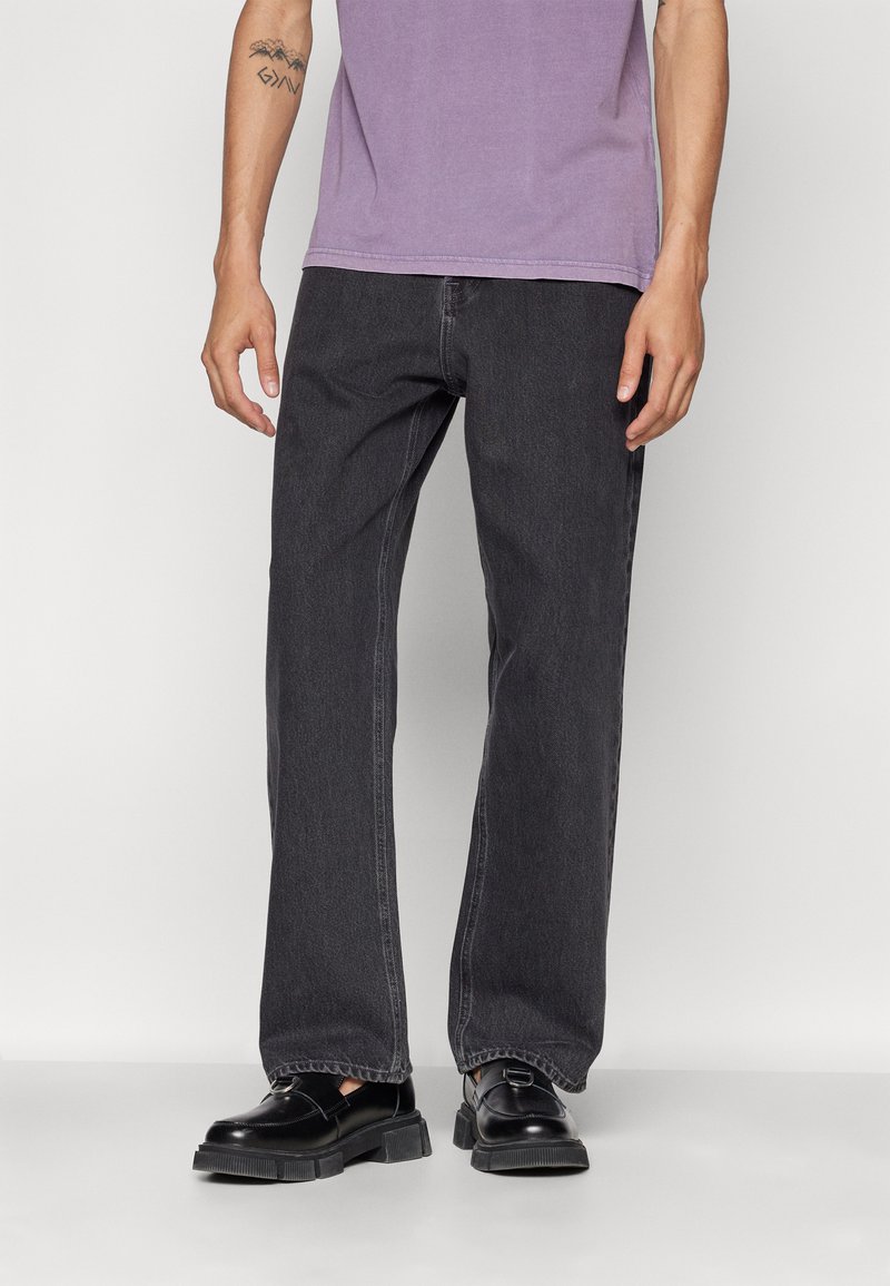 Jeans gris oscuro de corte holgado, hechos de denim, con piernas rectas y costuras sutiles. Llevados con una camiseta morada y zapatos negros.