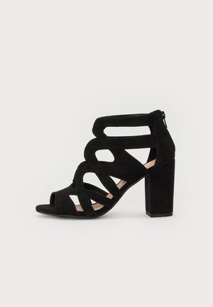 Zwarte suède hoge haksandalen met uitsnijdingen, open tenen en een blokhak. Ritsluiting aan de achterkant.