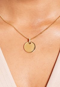 Gouden ketting met een ronde hanger met een gepolijste oppervlakte en een decoratieve schakel, rustend op blote huid.