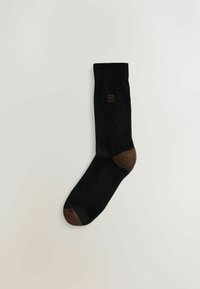 Schwarze Socke mit brauner Spitze und Ferse, mit einem kleinen braunen Logo an der Seite. Das Material wirkt glatt und dehnbar.