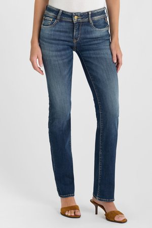 Donna che indossa jeans blu aderenti a vita media e sandali marroni con tacco e punta aperta, in piedi davanti a uno sfondo chiaro uniforme.