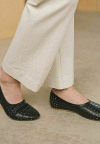 Bailarinas negras perforadas con un diseño de punta redonda, combinadas con pantalones de pierna ancha crema. Los zapatos cuentan con patrones recortados y una textura suave.