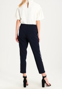 Kremowa bluzka z bufkami i dekoltem w kształcie łezki, w połączeniu z granatowymi spodniami o tapered fit z białymi paskami po bokach i sandałami na obcasie z paskiem przy kostce.