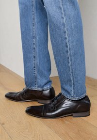 Chaussures habillées en cuir marron foncé au design élégant, bout arrondi et lacets minimalistes. Portées avec un jean en denim bleu.