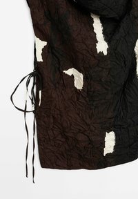 Tissu noir et marron froissé avec des taches blanches irrégulières et de fines attaches noires sur le côté gauche sur fond blanc.