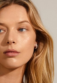 Primer plano de un rostro con cabello rubio largo y ondulado, tono de piel claro y un pequeño aro de pendiente con superficie texturada y brillante.