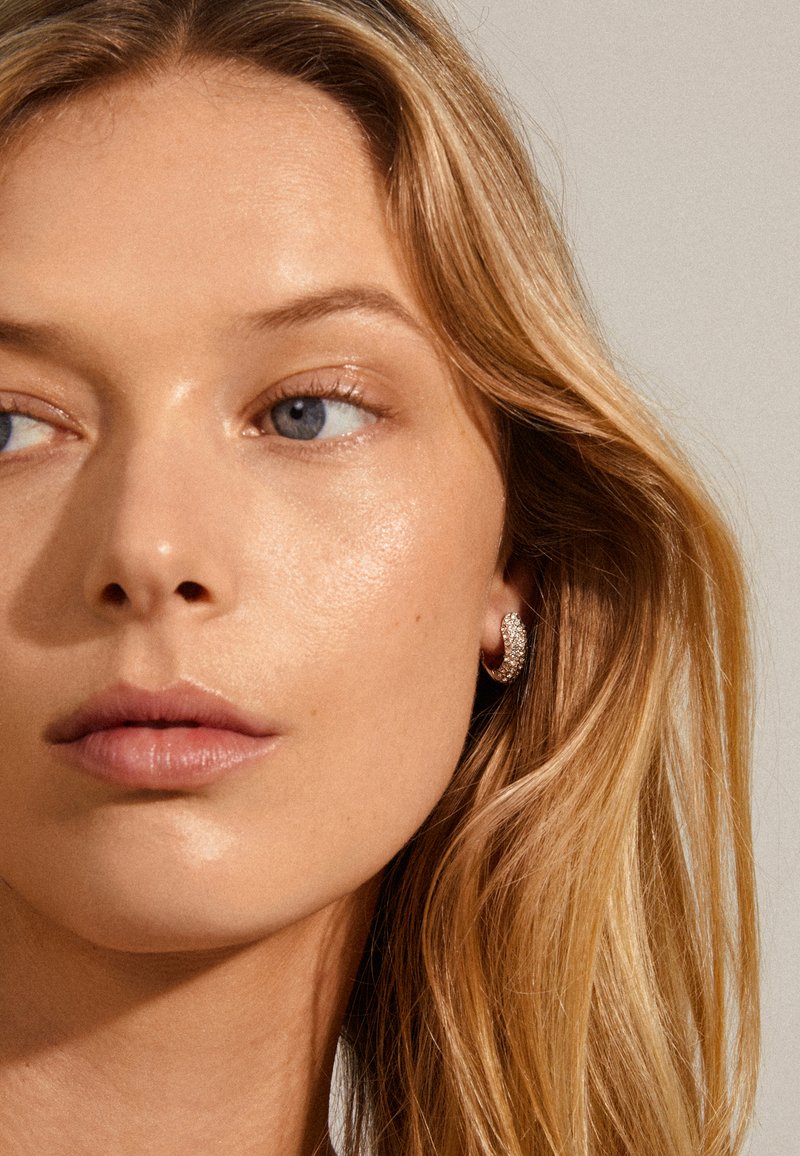 Primer plano de un rostro con cabello rubio largo y ondulado, tono de piel claro y un pequeño aro de pendiente con superficie texturada y brillante.