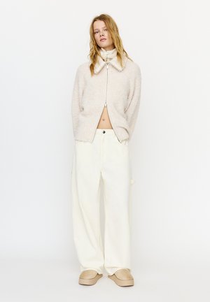 Młoda kobieta z rozpuszczonym blond włosami, ubrana w beżowy, miękki sweter na zamek, szerokie kremowe spodnie i beżowe buty wsuwane, stojąca na białym tle.