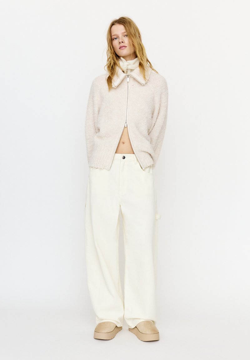 Młoda kobieta z rozpuszczonym blond włosami, ubrana w beżowy, miękki sweter na zamek, szerokie kremowe spodnie i beżowe buty wsuwane, stojąca na białym tle.