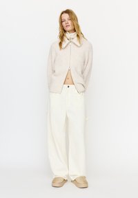 Jonge vrouw met los blond haar, gekleed in een beige zachte trui met ritssluiting, brede crèmekleurige broek en beige instapschoenen, staand tegen een witte achtergrond.