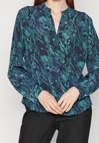 Blouse à manches longues en bleu foncé avec un motif abstrait en turquoise tourbillonnant. Dotée d'un col mandarin et de détails de poignets à boutons.